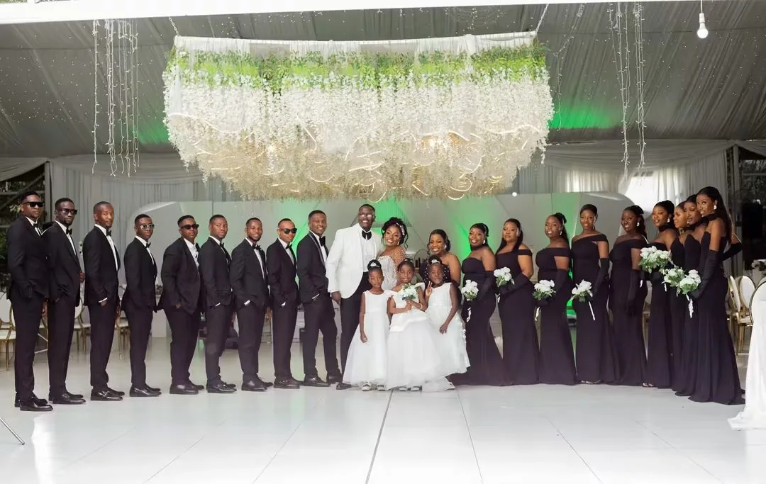 Bridal party in black gowns and tuxedos under the floral chandelier — founder's pre-Caronai experience, planned and coordinated by Tanaka
